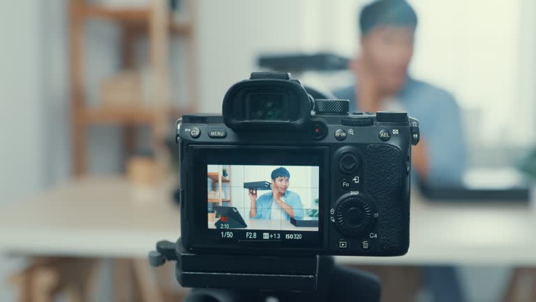 Close up of Young Asian man blogger opening parcel box and recording unboxing video with camera sitting at work desk in living room at home.