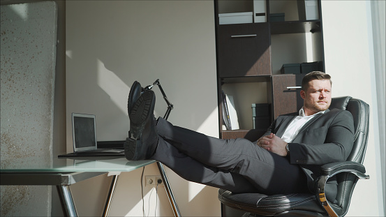 Exhausted businessman sits with his feet on his desk and thinks about the crisis. A businessman in his office throws his feet on the table.