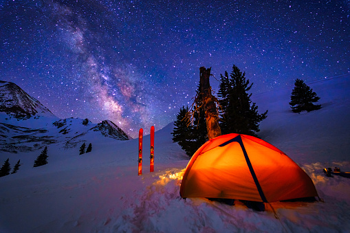Ski Mountaineering Winter Camping - Ski camp setup with glowing tent at high altitude under mountain ridge with stars and Milky Way Galaxy.