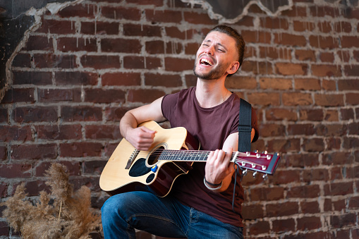 Man with acoustic guitar leaning brick wall playing music singing songs enjoy life Handsome caucasian male guitar player practice play musical instrument home loft interior Creative lifestyle