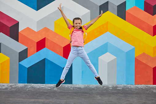 Full body of carefree schoolgirl with backpack jumping with raised arms on colorful background and looking at camera
