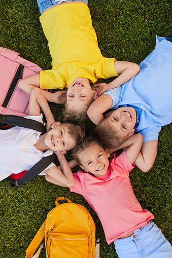 Top view of group of delighted schoolchildren with rucksacks lying on green meadow and looking at camera in park