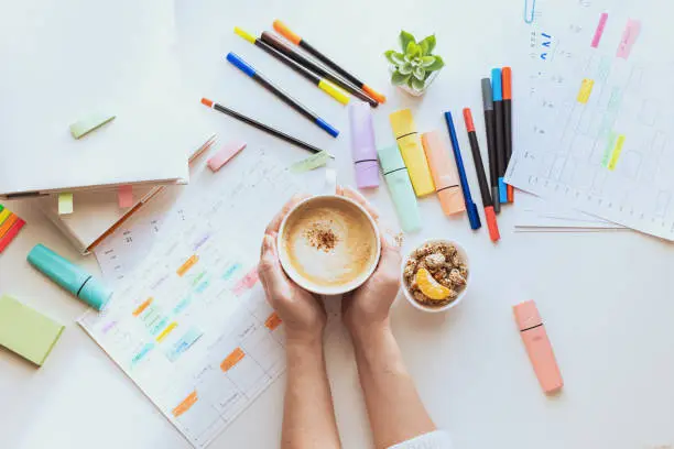 Photo of Student planning process. The student's desk with a weekly schedule, books, colorful markers and hands with a cup of coffee. View from above.