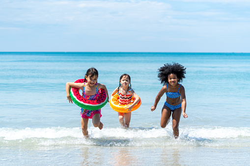 Little African and Asian girl in swimwear and swim ring playing in sea water together at tropical beach in sunny day. Happy children enjoy and fun outdoor activity lifestyle on summer holiday vacation