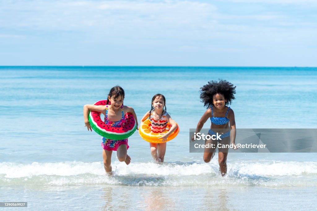 Little African and Asian girl playing in sea water together at tropical beach on summer vacation Little African and Asian girl in swimwear and swim ring playing in sea water together at tropical beach in sunny day. Happy children enjoy and fun outdoor activity lifestyle on summer holiday vacation Beach Stock Photo