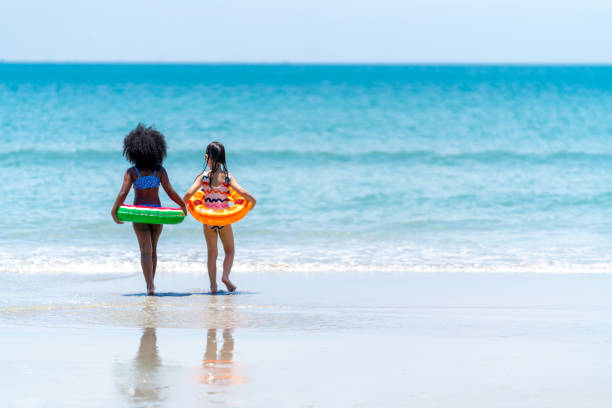 Little African and Asian girl playing in sea water together at tropical beach on summer vacation Little African and Asian girl in swimwear and swim ring playing in sea water together at tropical beach in sunny day. Happy children enjoy and fun outdoor activity lifestyle on summer holiday vacation family beach vacations travel stock pictures, royalty-free photos & images