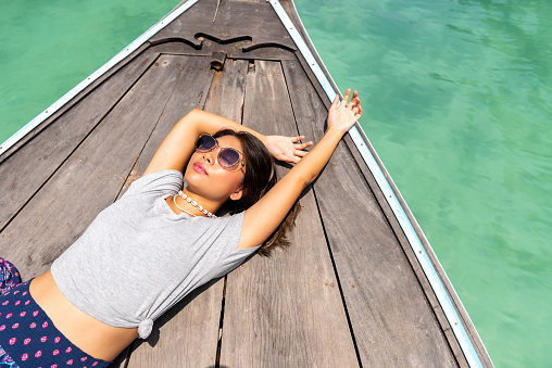Portrait of Young beautiful Asian woman lying on the boat passing beach lagoon in summer sunny day. Happy female relax and enjoy outdoor lifestyle on holiday travel vacation at tropical island in Thailand
