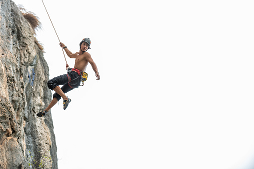 Confidence young Asian man climber with safety equipment climbing on rocky mountain at tropical island in sunny day. Strong handsome male enjoy outdoor active lifestyle and extreme sport on summer vacation