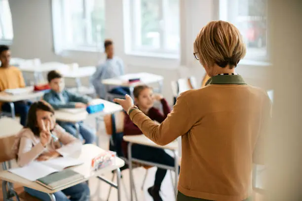 Rear view of elementary school teacher pointing at one of her students who is raising arm to answer a question.