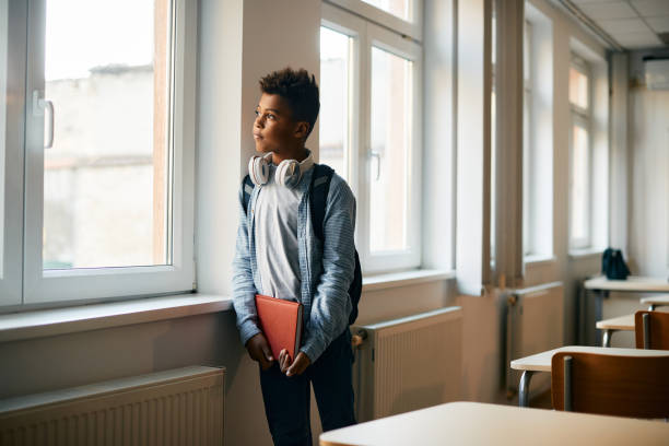 Pensive black schoolboy looking through the window in the classroom.