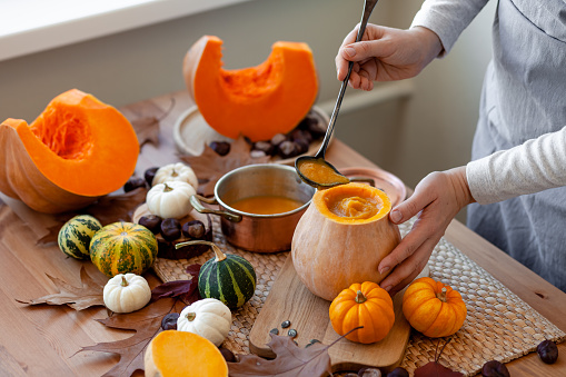 Young woman in apron preparing delicious homemade pumpkin soup on her kitchen. Fresh organic ingredients, traditional autumn food. Healthy eating, cozy home atmosphere