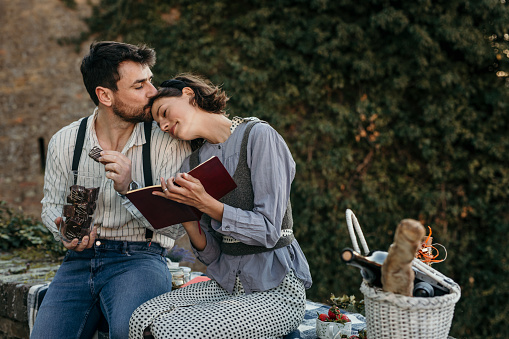 Newly weds enjoying their picnic time in a fortress, sitting on a brick wall, both retro urban stylishly dressed. A cute bearded guy is eating chocolate cube while enjoying the story that his loving wife, a brunet, is reading to them, leaning on his chest.