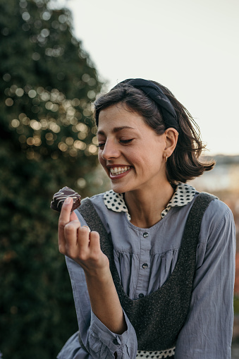 Cheerful female sitting on a balcony in a retro dress and a headband on her head, holding a chocolate dessert and about to eat it. People and sweets concept