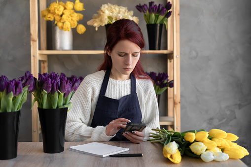 a young female florist in a blue apron checks orders on a mobile phone while sitting at a table with fresh flowers in glass vases in a store. copy space.