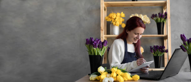 portrait of a florist in an apron, working in her own flower shop, using a laptop organizing logistics and delivery, taking orders online, taking notes with a pen in a notebook. banner. - flower computer young women selling imagens e fotografias de stock