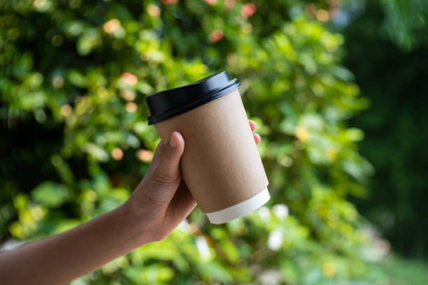 hand holding paper cup of take away drinking coffee on green trees in the background.