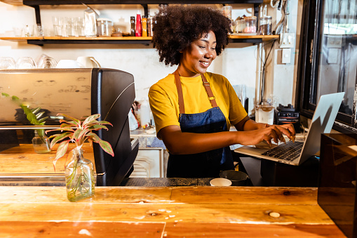 Young Female manager in restaurant with digital tablet or notebook.Close up of joyful African American young woman worker in apron stands in cafe restaurant.Small business concept.