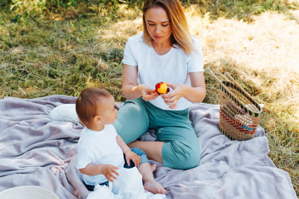 Happy family picnic in nature, mom and baby are resting on a blanket on the green grass outside. Beautiful young mother cleaning fruit for her toddler son, outdoors. stock photo