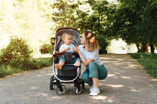 Family portrait, young fashionable mother holding the hand of her little child sitting in a stroller, outdoors. Mom and son on a summer walk, lifestyle. Family and motherhood concept.