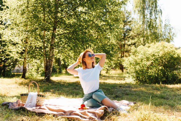 Laughing young woman in glasses and a hat on picnic in nature. Smiling beautiful girl sitting on blanket on the green grass with fruits and basket. Positive woman enjoying summer vacation, outdoors. stock photo