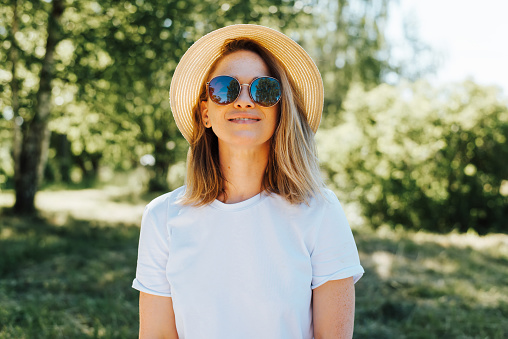 Portrait smiling pretty freckled young woman in straw hat and sunglasses looking away on summer sunny day outdoors. Cute caucasian girl in white t-shirt in nature.