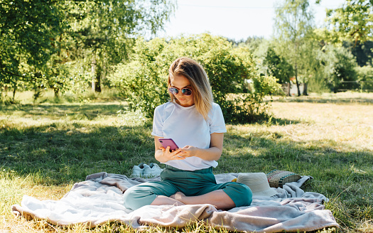 Young woman browsing the Internet on a smartphone while relaxing in nature on a summer day. Beautiful girl in sunglasses sitting on a blanket on the lawn and using a mobile phone, outdoors.