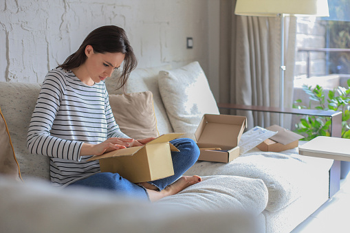 Beautiful young woman is holding cardboard box and unpacking it sitting on sofa at home