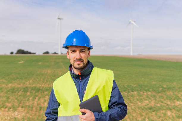 retrato de um jovem engenheiro industrial em um parque de turbinas eólicas usando seu tablet digital. trabalhador olhando para a câmera nas instalações de energia elétrica renovável e não poluente. - fuel and power generation nonpolluting wind turbine - fotografias e filmes do acervo