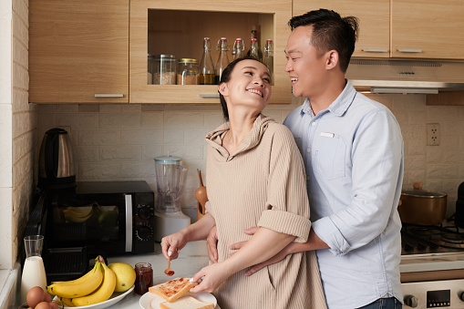 Happy Vietnamese man hugging wife making jam toasts for breakfast