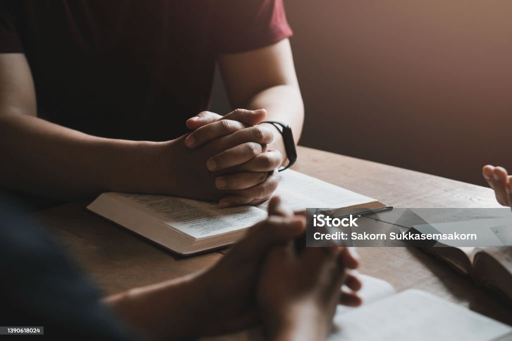 A group of Christians sit together and pray around a wooden table with blurred open Bible pages in their homeroom. Prayer for brothers, faith, hope, love, prayer meeting Minister - Clergy Stock Photo