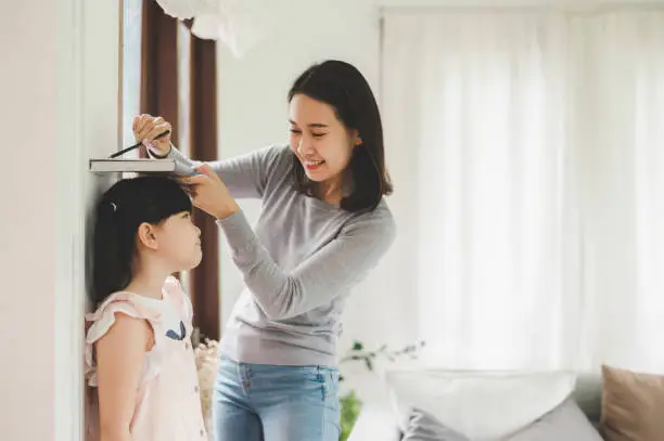 Happy Asian mother using book and pencil to measure height of little girl near wall at home