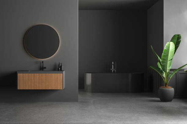 Dark bathroom interior with concrete floor, black bathtub, and oval mirror standing on black wall.
