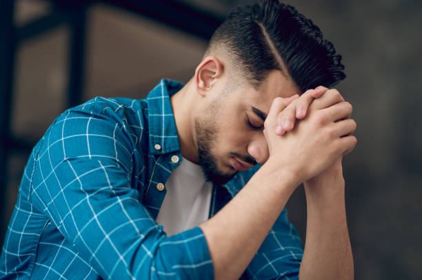 Young dark-haired man sitting with an unhappy look Stressed. Young dark-haired man sitting with an unhappy look prayer stock pictures, royalty-free photos & images