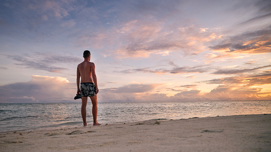 Rear view of young man on idyllic white sand beach. Tourist in swimwear watching colorful sunset in Maldives.