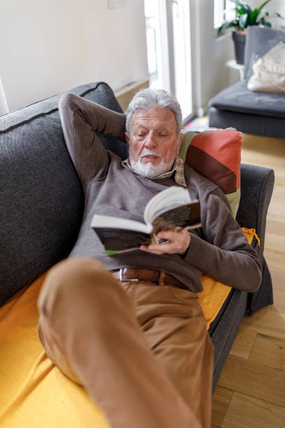 istock Handsome bearded senior man reading book on the sofa at home. 1390650685