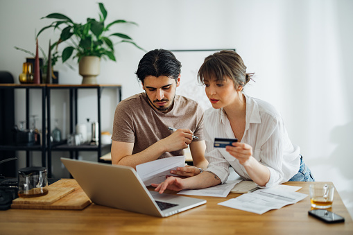 Boyfriend looking at bills while his girlfriend holding credit card and talking with him while doing home finances together online on a laptop computer in the kitchen.