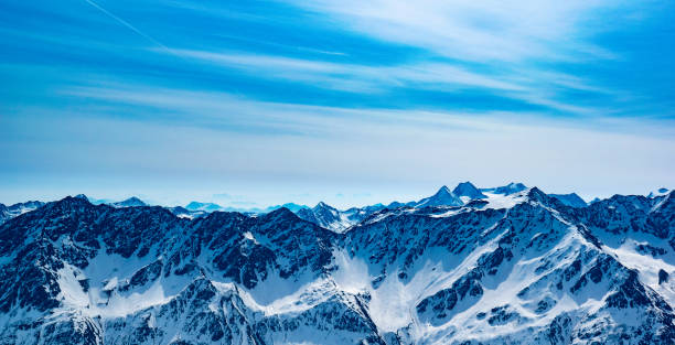Thin striations of cloud hover above the Austrian alps, which stretch off into the horizon. The rock and snow catch the light and shadows cast by the Springtime sun, which was high in the sky.  Numerous peaks break into the horizon beyond, demonstrating the scale of this majestic alpine environment.