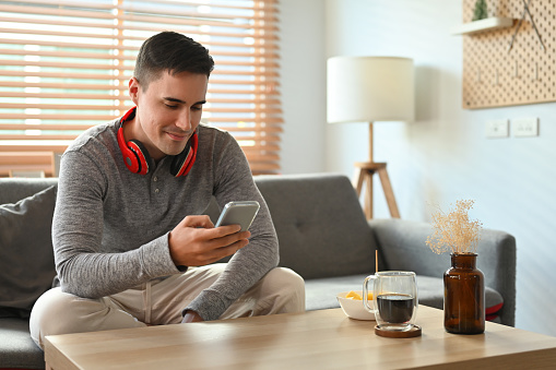 Handsome Caucasian man on a grey shirt sitting on a couch at home using a smartphone.