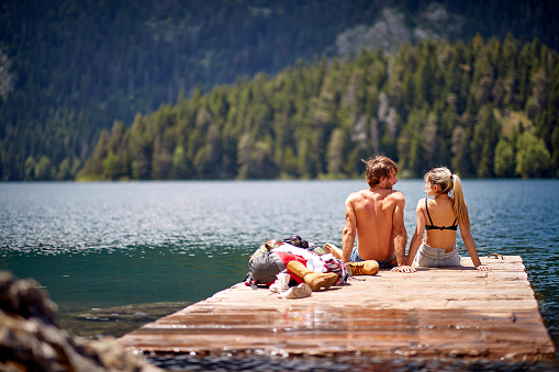 A young couple is taking a sunbath on the dock at the lake during mountain hiking on a beautiful day. Trip, nature, hiking