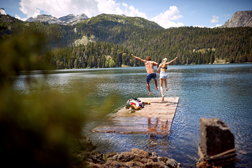 A young couple is jumping into the lake during mountain hiking on a beautiful day. Trip, nature, hiking concept