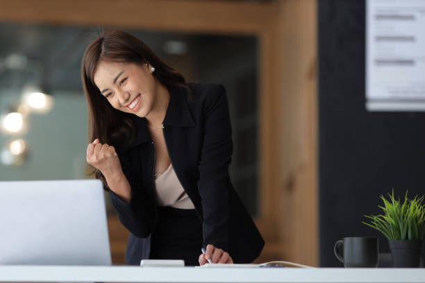 seorang wanita bisnis atau sekretaris yang bekerja di laptop mengungkapkan kegembiraannya sambil melihat layar laptop. karyawan wanita asia mengekspresikan kegembiraan kesuksesan dan bonus. - sukses potret stok, foto, & gambar bebas royalti