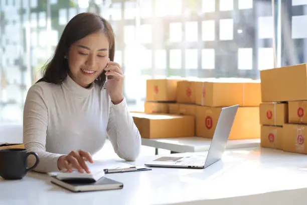 Photo of A young woman, e-commerce online small start-up business owner, calls with a partner and uses a calculator to calculate the cost and shipping costs.