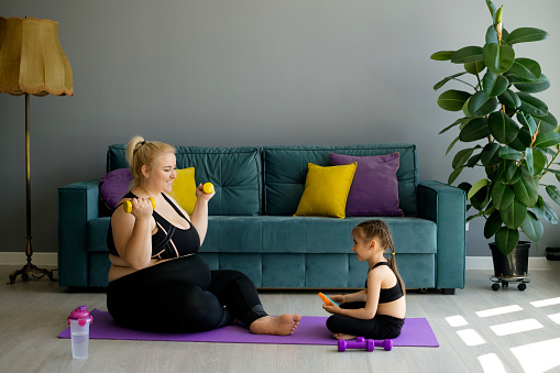 A fat obesity plump pregnant mother goes in for sports with little daughter, who provides moral support to mother. Joint fitness training with dumbbells. Person lady trains at home in the hallway on the floor