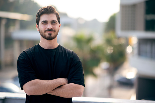 Portrait of happy young man in casual clothing looking at camera outdoor. Smiling man with beard feeling confident. Successful business man in city street.
