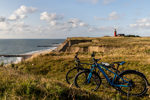Paraglider soaring along the cliffs of Bovbjerg, Denmark. Scenic view from top of the cliffs onto the sea, the lighthouse Bovbjerg Fyr in the background, two bicycles in front