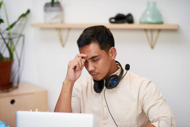 A young Asian businessman rubs his eyes at his desk in the office looking upset stock photo