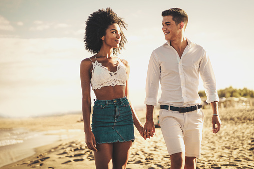 Gen z biracial couple of newlyweds on vacation on the beach walking hand by hand and looking each other eyes