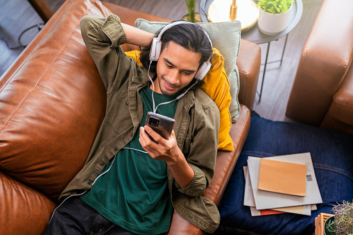 Asian man in headphones listening to the music and gesturing while sitting on his couch.clam and peaceful at home cheerful asian young male with a headset and smartphone playlists choose to playing songs on the smartphone in the living room at home