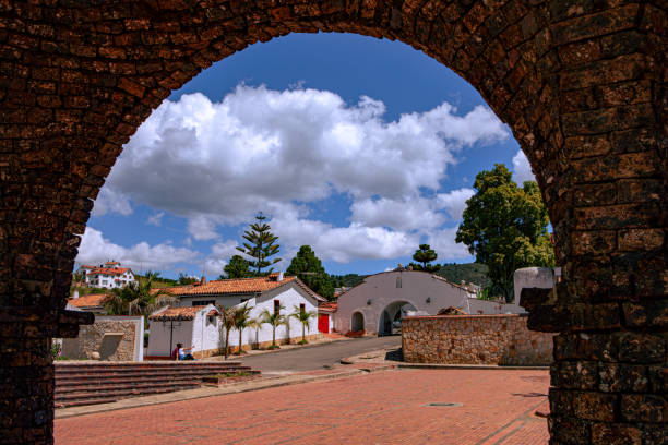 Guatavita Town, Colombia, South America - Looking from under an archway of the principal entrance to the main Town Square, towards the gateway to the square in the Andes town, in the Cundinamarca Department of the South American country of Colombia. Though completed in the 1960s, the town is designed and built in the Spanish colonial style of architecture. Situated about an hour's drive from the capital city of Bogotá, it is located at an altitide of 8,660 feet above mean sea level and is a popular weekend destination with both local Colombians, and tourists. Photo shot in the afternoon sunlight; horizontal format. Copy space. Note to inspector: People's faces have been blurred in Photoshop and cannot be recognised. mirando hacia el centro de terracota casas con techo de tejas en la ciudad andina de guatavita en colombia, américa del sur, a través de una puerta de entrada arqueada en la plaza principal de la ciudad. arquitectura de estilo colonial español.
