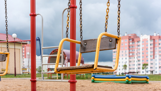 old brown yellow swing in local playground with attractions against blurry city buildings under cloudy sky close view